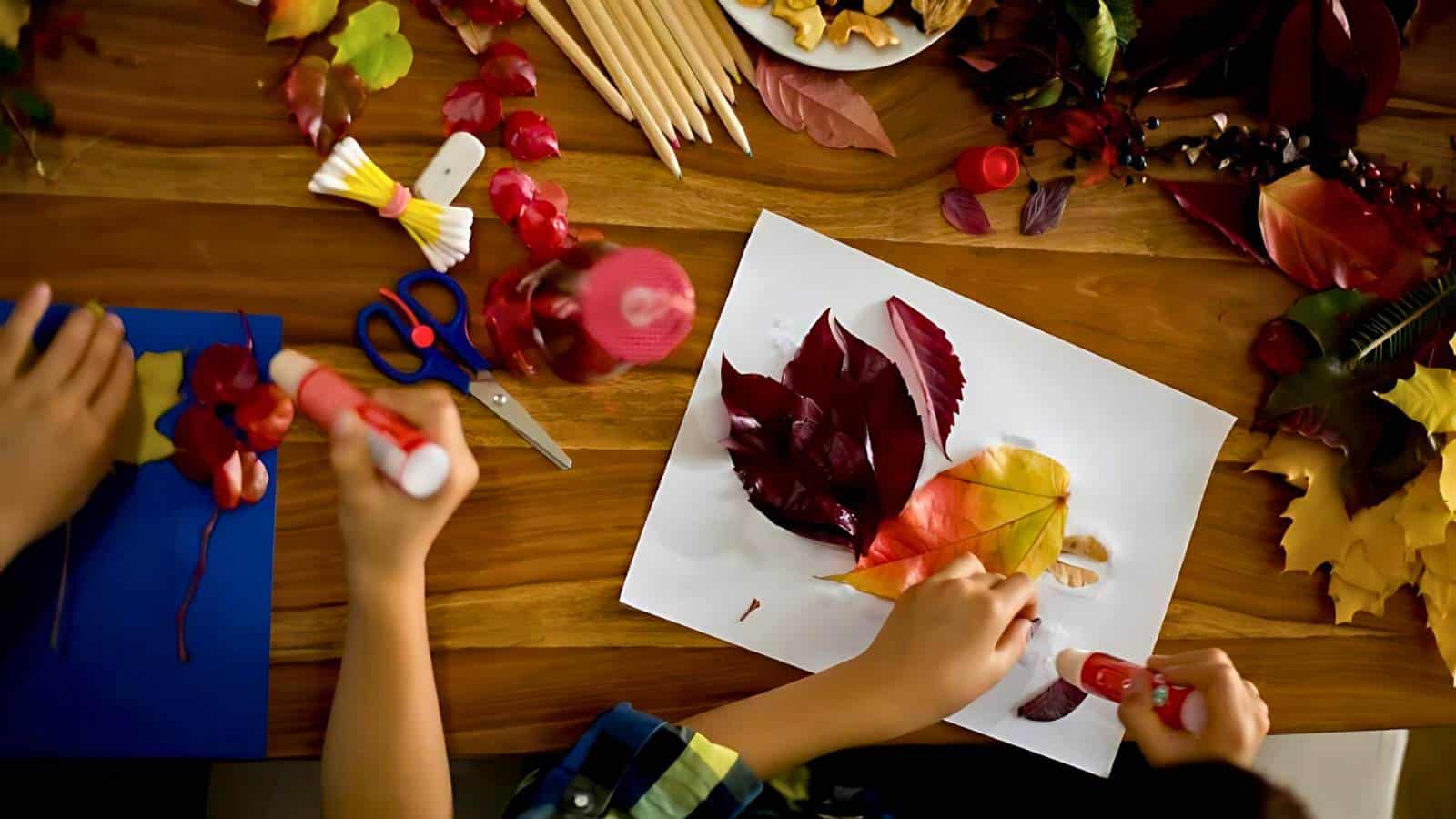 Two children creating autumn leaf crafts on a wooden table, using glue, scissors, and paper.