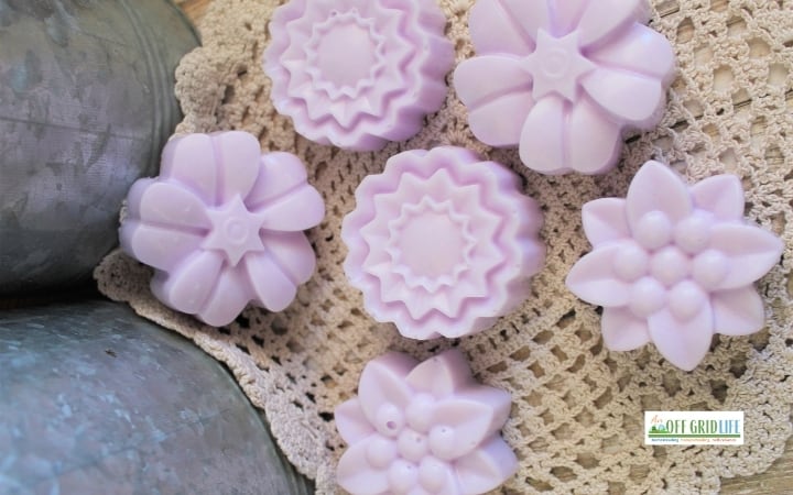 Six lavender-colored flower-shaped soap bars, inspired by whipped sugar scrub soap recipes, are arranged on a crocheted doily beside a metal surface. A small "Off Grid Life" logo appears in the lower right corner.