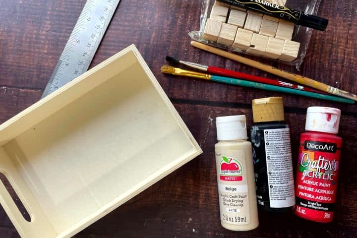 A wooden box lies next to a ruler, paint brushes, acrylic paints, wooden blocks, and paint markers on a dark wooden surface.