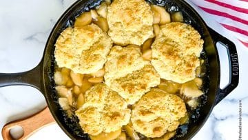 A skillet of apple cobbler with six biscuit toppings on a marble surface.