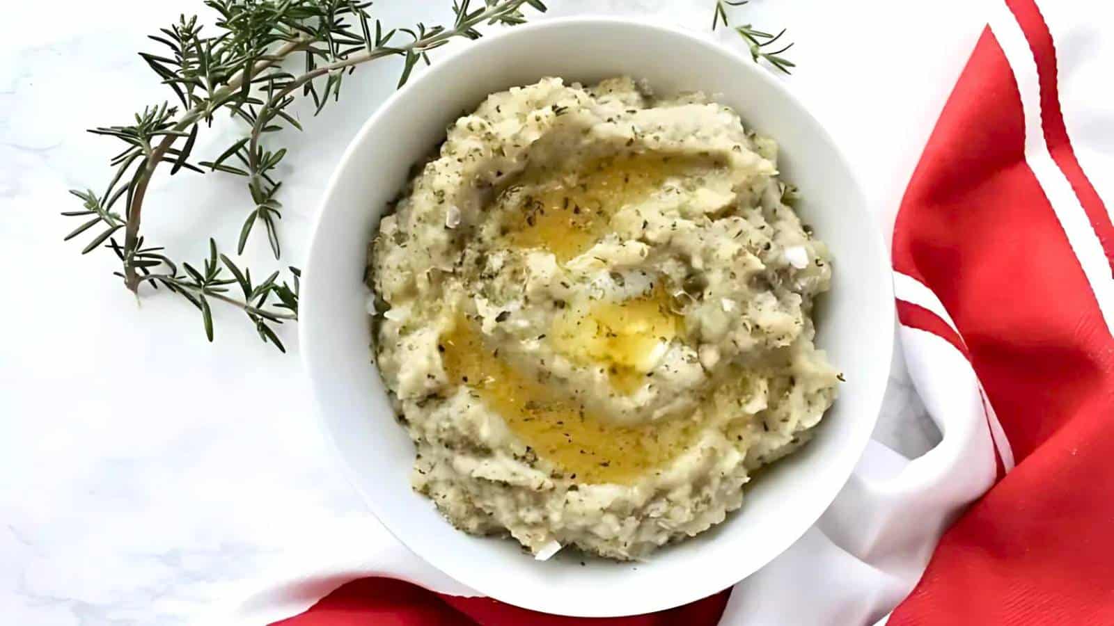 A bowl of mashed potatoes topped with herbs and a drizzle of melted butter sits beside a sprig of rosemary and a red and white cloth.