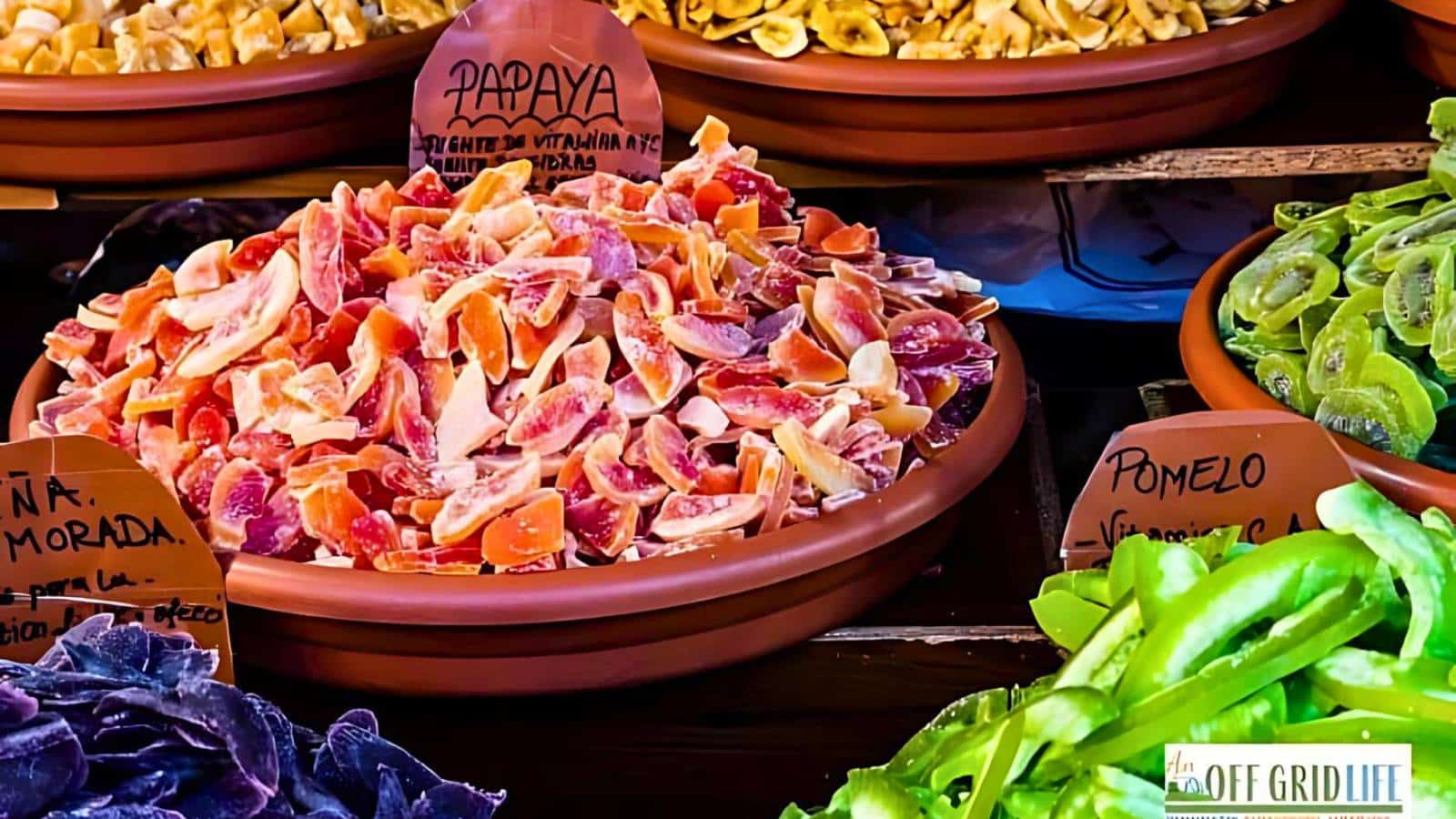 A market display featuring bowls of various dried fruits, including a bowl of dried papaya labeled "Papaya" and other visible fruits such as dried pomelo, with green and purple fruits nearby.