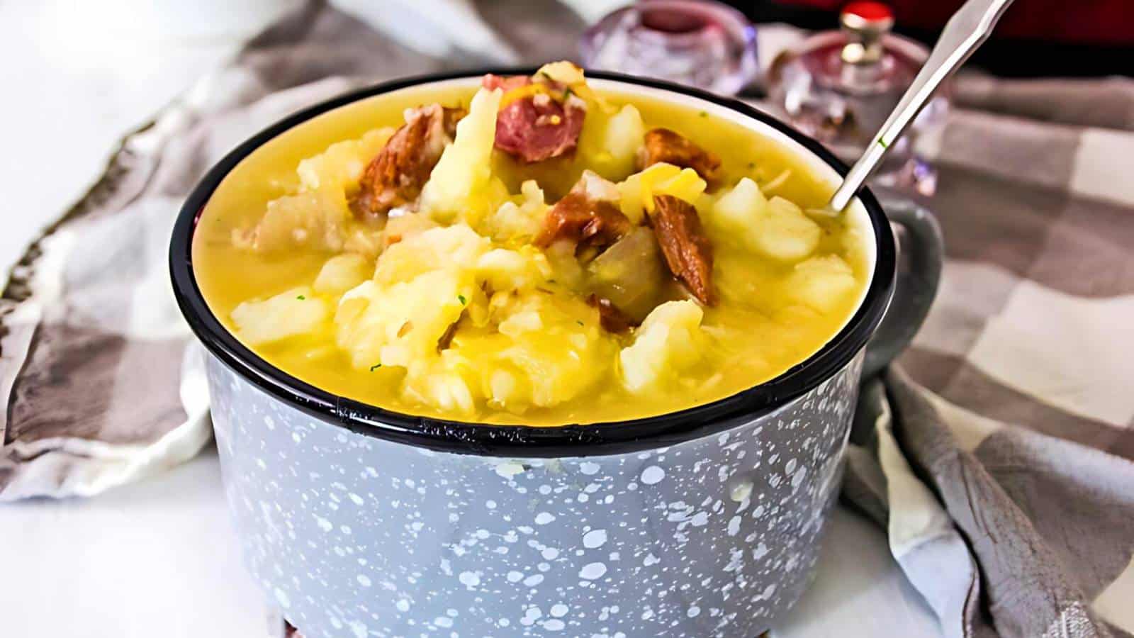A speckled gray bowl filled with a thick potato soup containing meat chunks, with a spoon inside the bowl, placed on a checkered cloth.