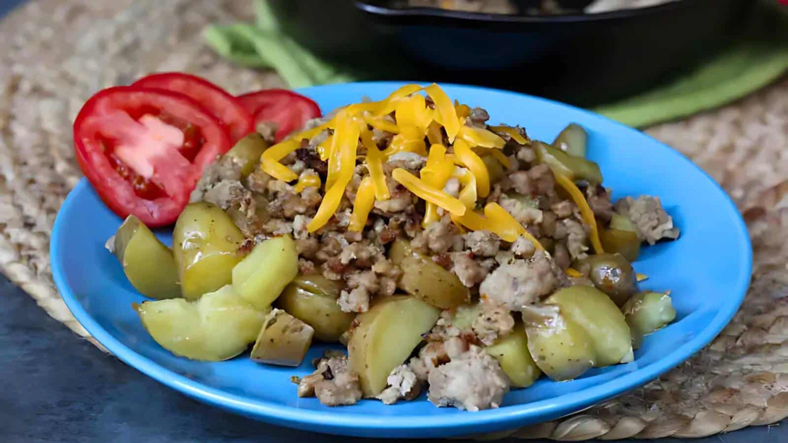 A plate of diced cooked squash with ground meat and shredded cheese, garnished with three slices of tomato, on a woven mat.