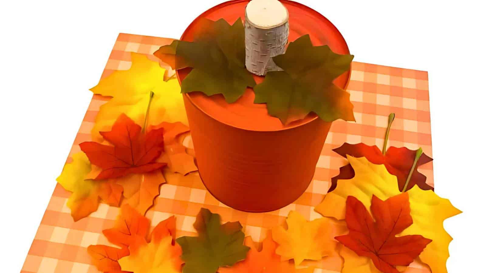 An orange container with a birch stem lid sits on a checkered surface, surrounded by various colorful autumn leaves.