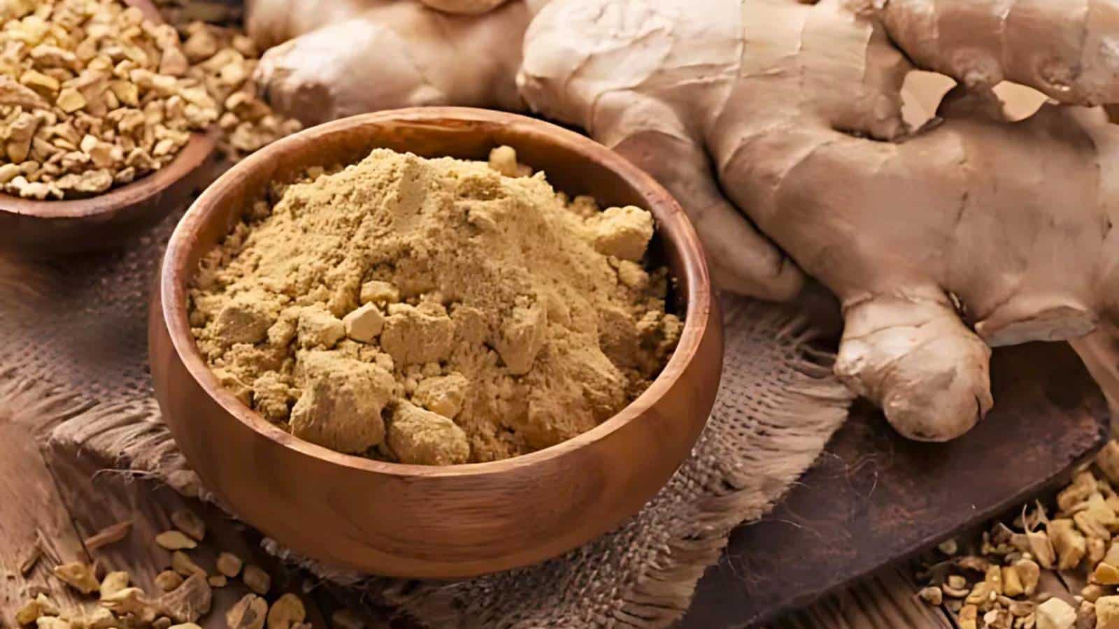 A bowl of yellow liquid on a wooden surface surrounded by scattered yellow flowers.