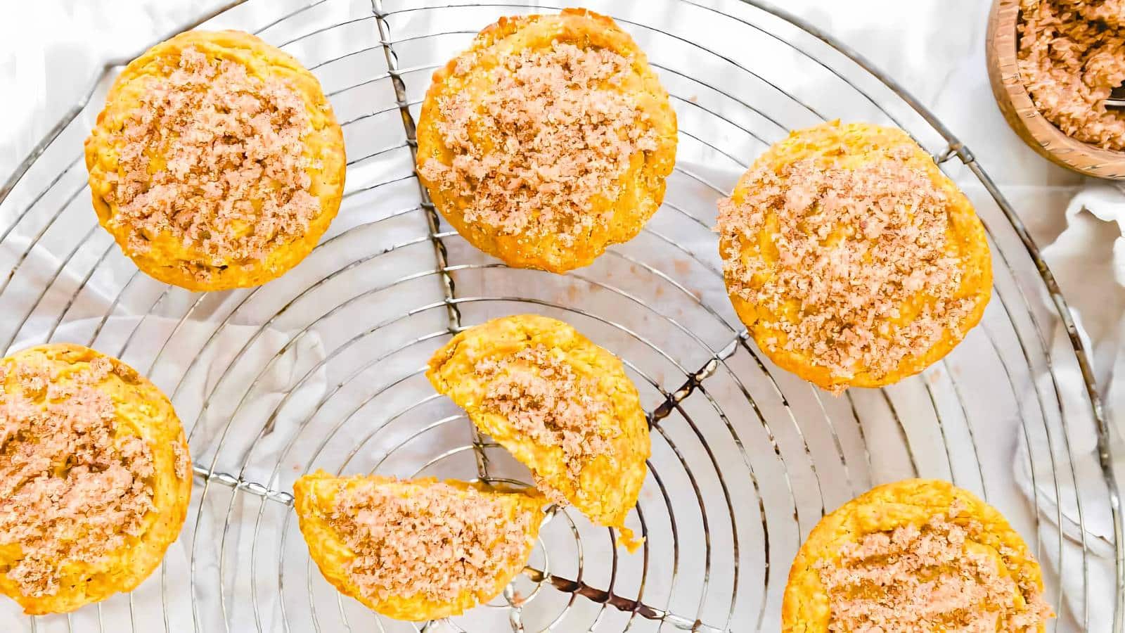 Six muffins topped with streusel are arranged on a round cooling rack, with one muffin cut in half. The background features a light-colored surface.