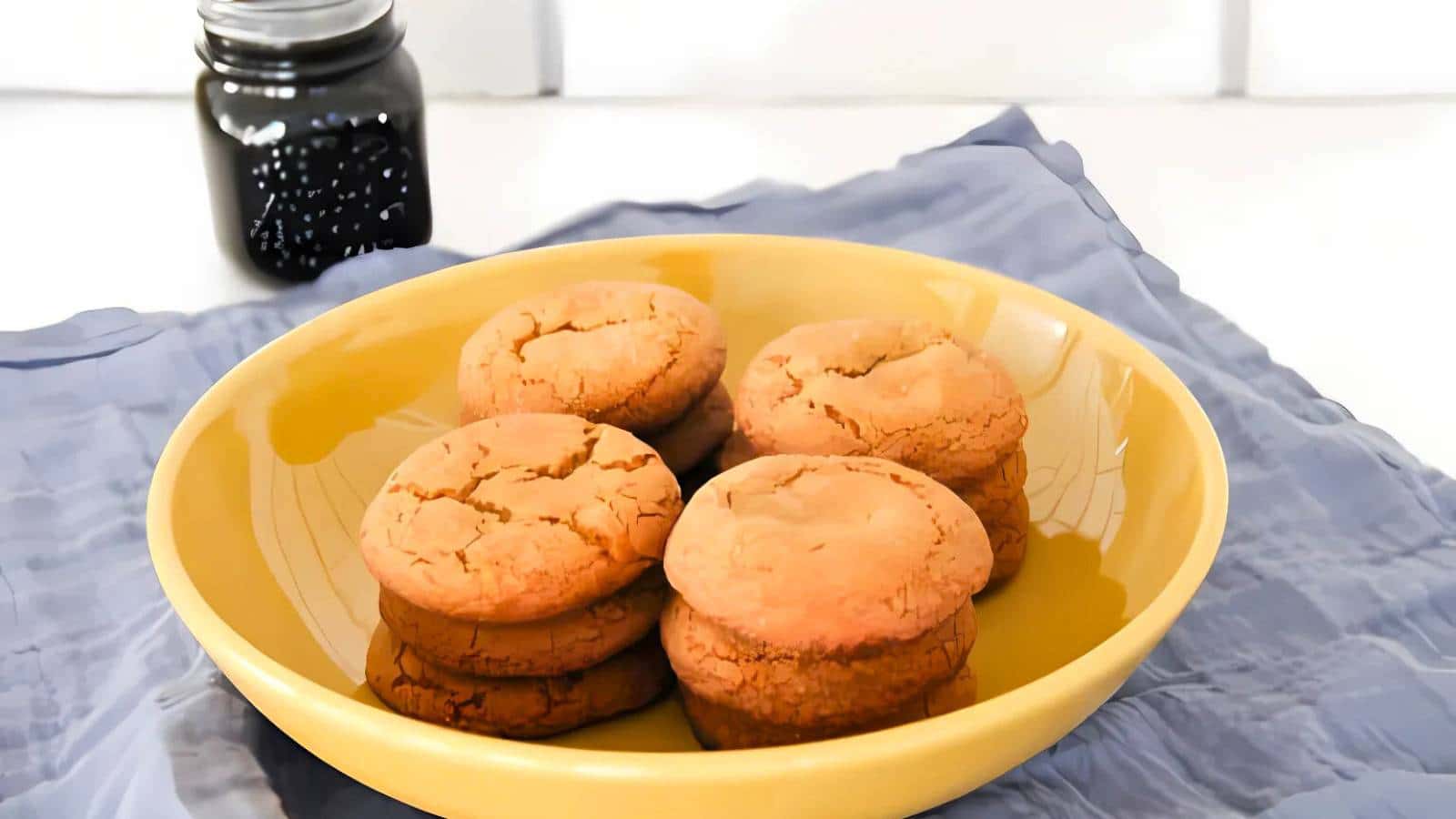 A yellow bowl holding a stack of six cookies and bars rests on a blue cloth, while a jar with a dark substance sits in the background.