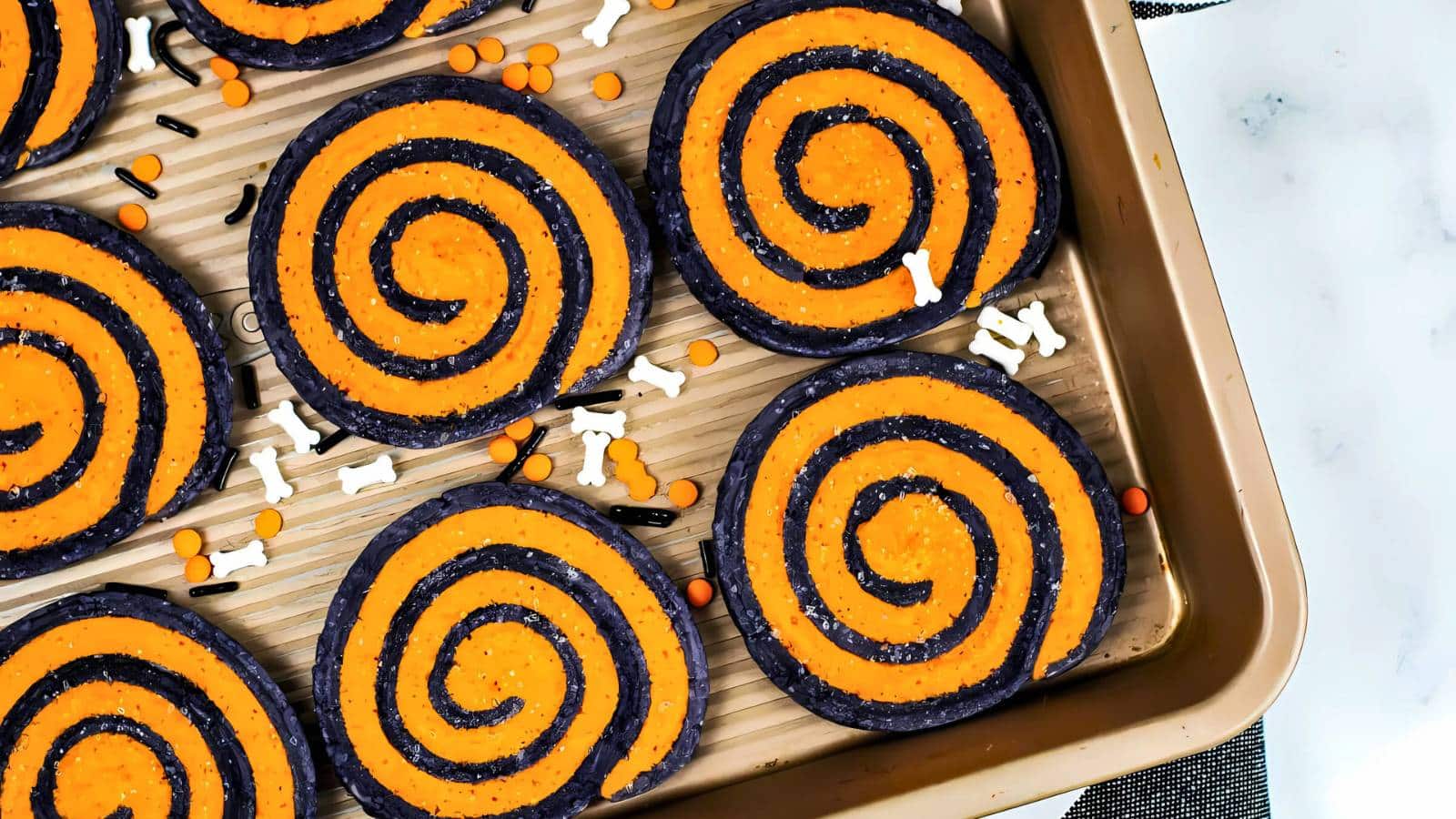 A baking tray with spiral cookies featuring orange and black swirls, decorated with bone-shaped and round sprinkles.