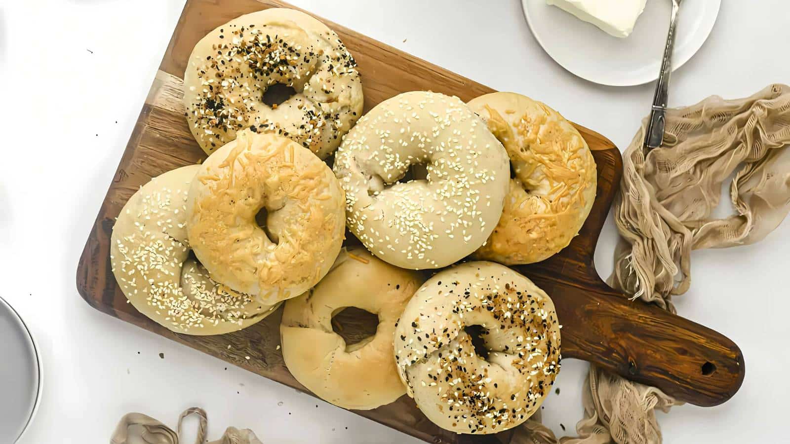 A wooden board with seven bagels, some topped with sesame seeds and others with everything seasoning, next to a knife with butter.