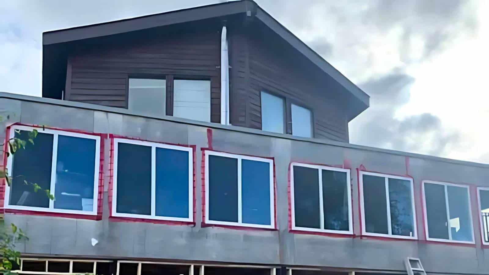 A two-story building with a wooden upper level and a lower level featuring large windows lined with red trim. Overcast sky in the background.
