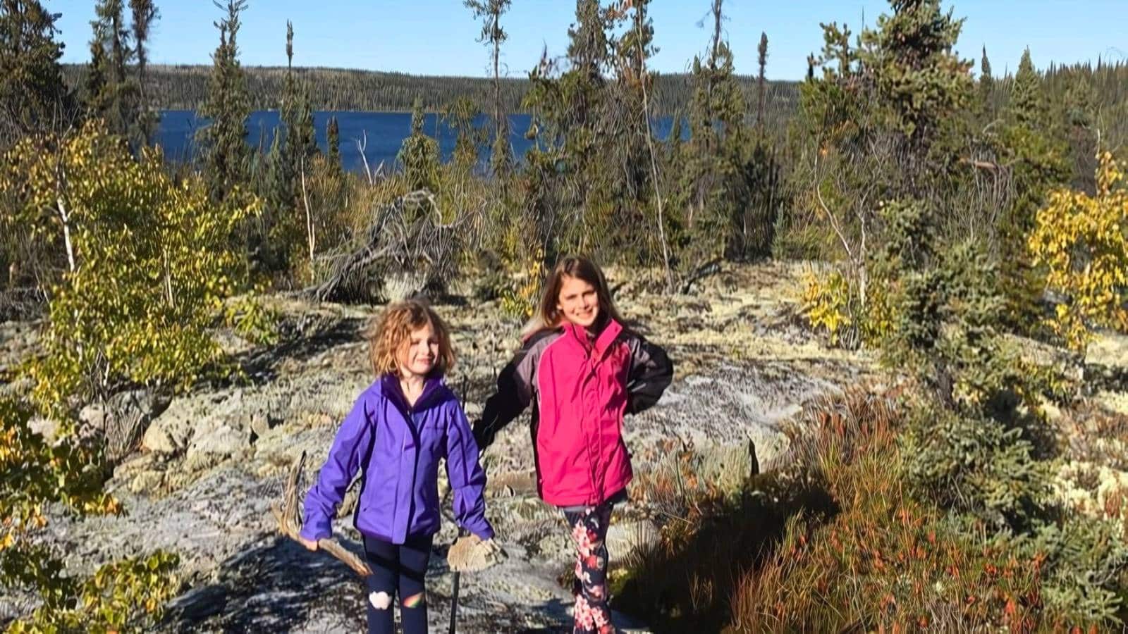 Two children stand on a rocky landscape, surrounded by trees with a lake in the background. They are wearing jackets, with one in purple and the other in pink.