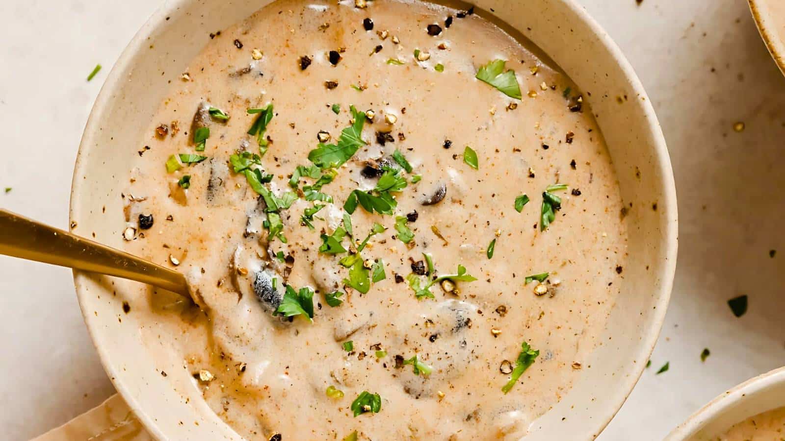A bowl of creamy mushroom soup garnished with chopped parsley and cracked black pepper. A spoon rests inside the bowl.