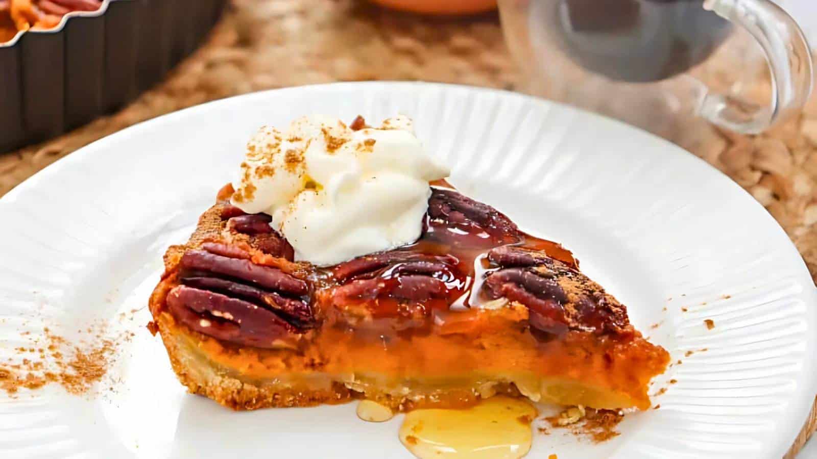 A slice of pecan pie on a white plate, topped with whipped cream and drizzled with syrup—a perfect example of yummy Thanksgiving desserts. A cup of coffee is in the background.