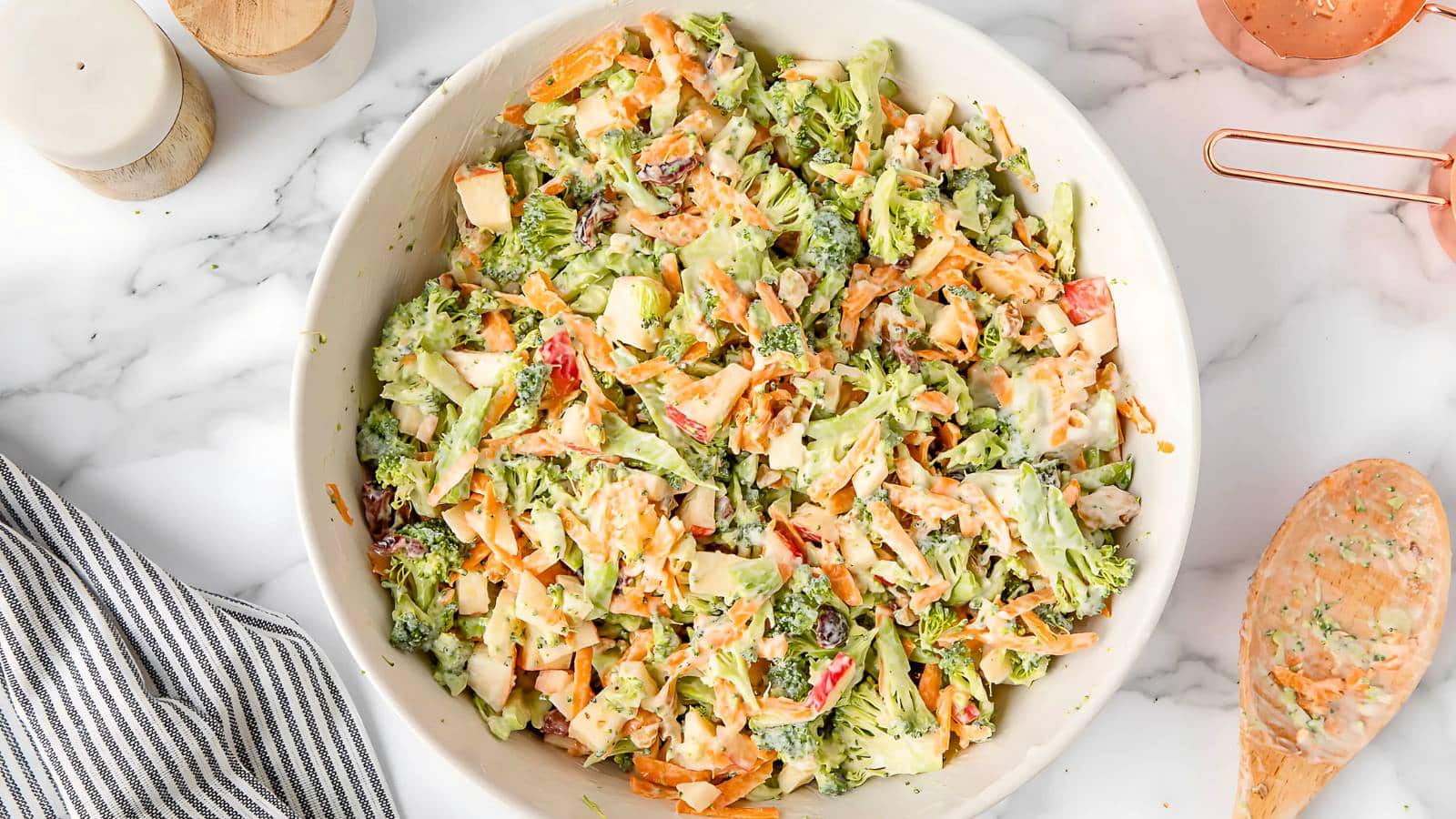 A bowl of broccoli salad with carrots, apples, raisins, and a creamy dressing on a marble surface next to a wooden spoon and striped cloth.