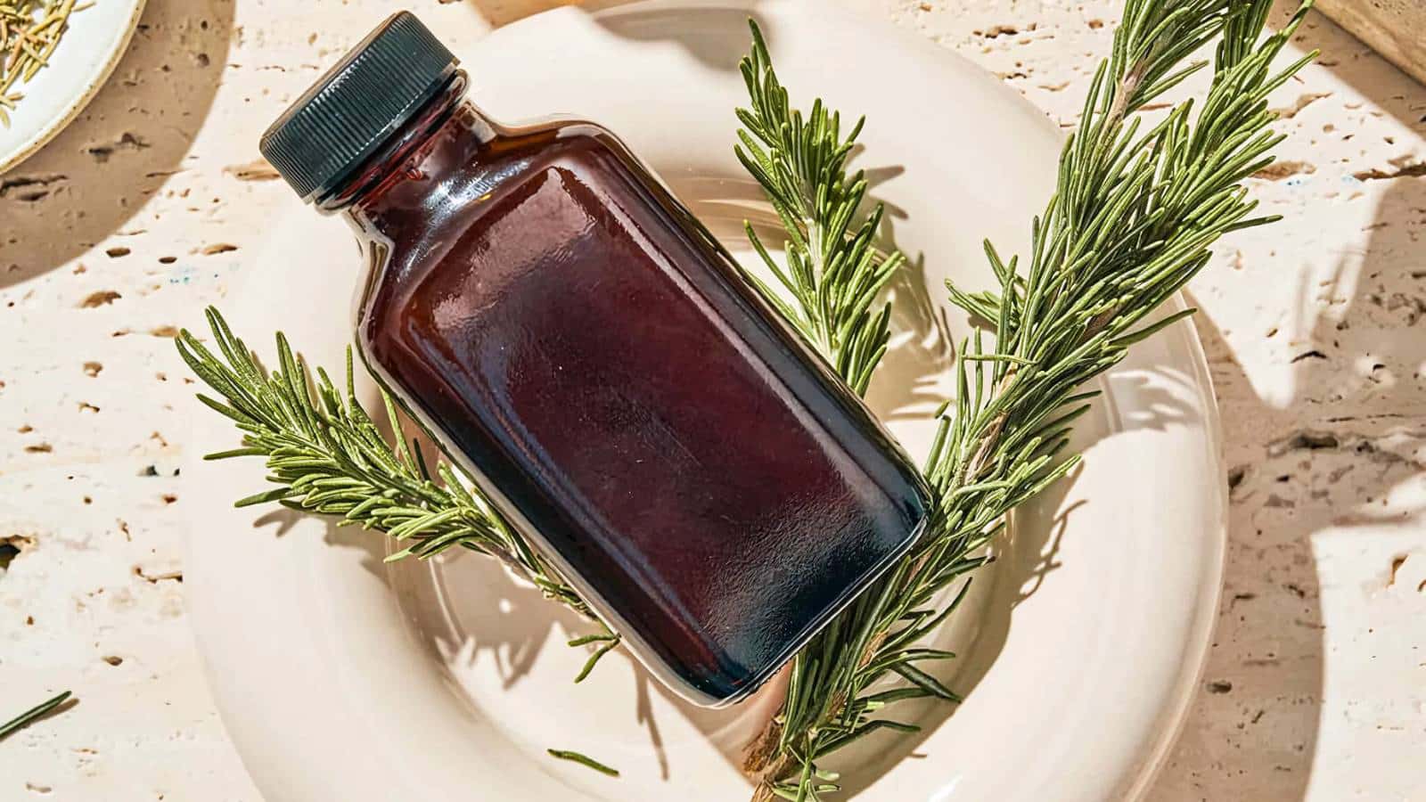A dark glass bottle with a black cap is placed on a white ceramic plate, surrounded by sprigs of fresh rosemary.