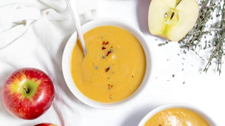 A bowl of creamy soup garnished with herbs next to red apples, apple halves, and sprigs of thyme on a white background.