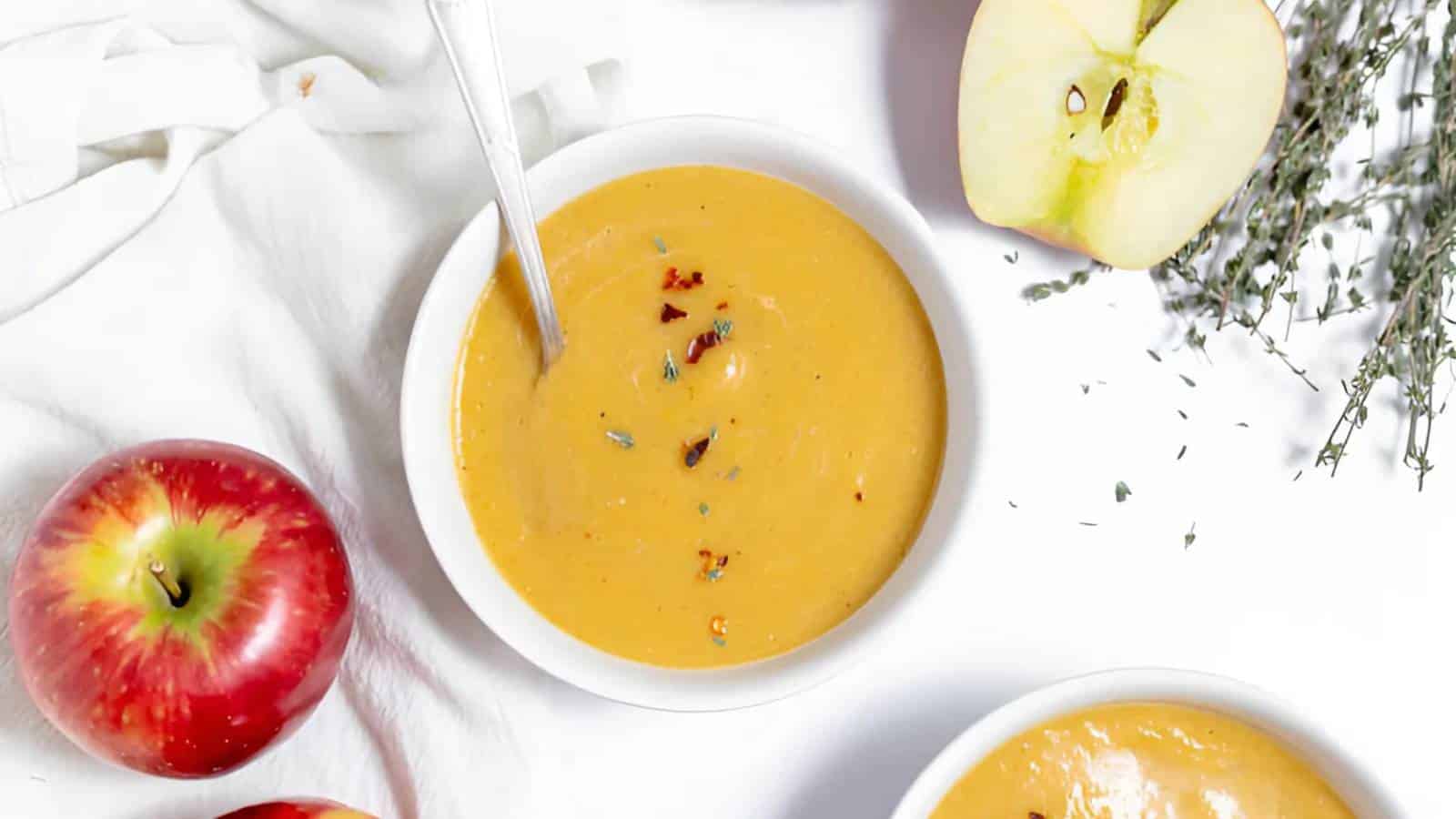 A bowl of creamy soup garnished with herbs next to red apples, apple halves, and sprigs of thyme on a white background.
