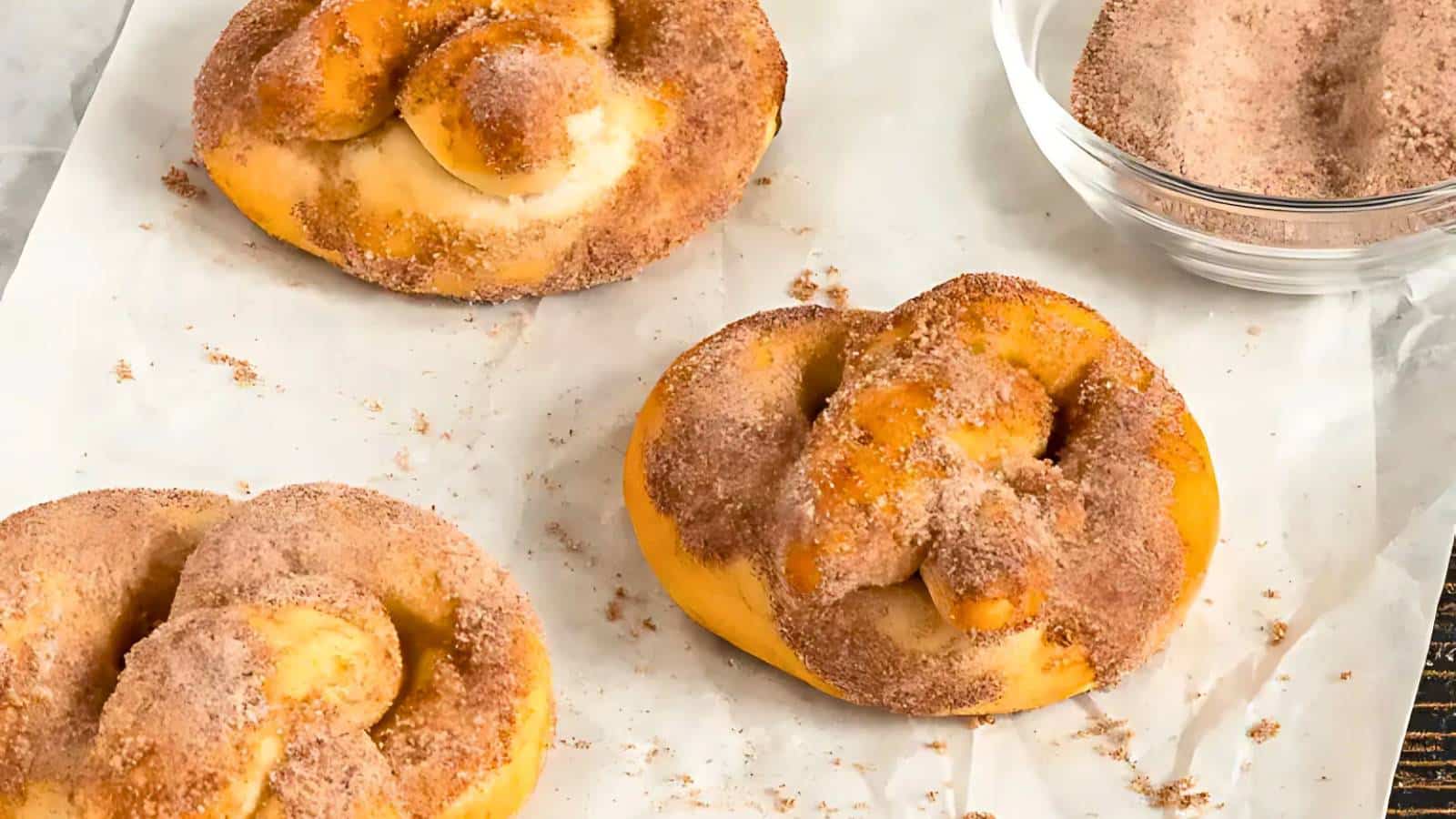 Three cinnamon sugar pretzels on parchment paper, with a bowl of cinnamon sugar mixture nearby.