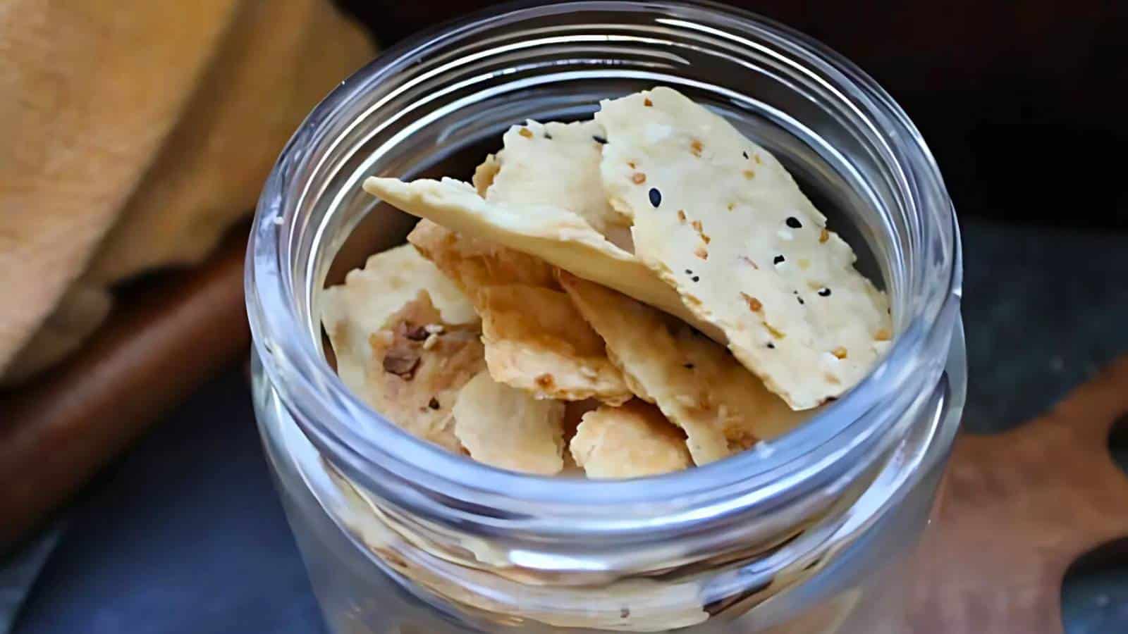 A glass jar filled with scattered flat crackers seasoned with sesame and spices, resting on a textured surface.