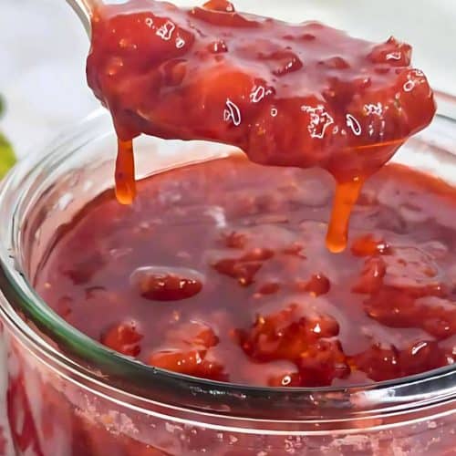 A close-up of a spoon lifting strawberry jam from a glass jar. A cluster of red flowers and green leaves is blurred in the background.