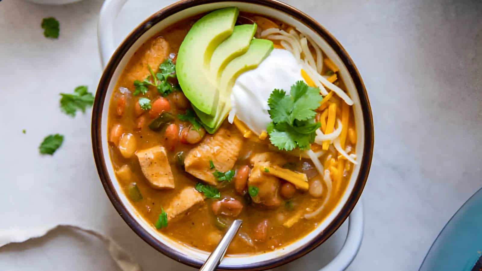 Bowl of chili with chicken, beans, cheese, sour cream, avocado slices, and a cilantro garnish.