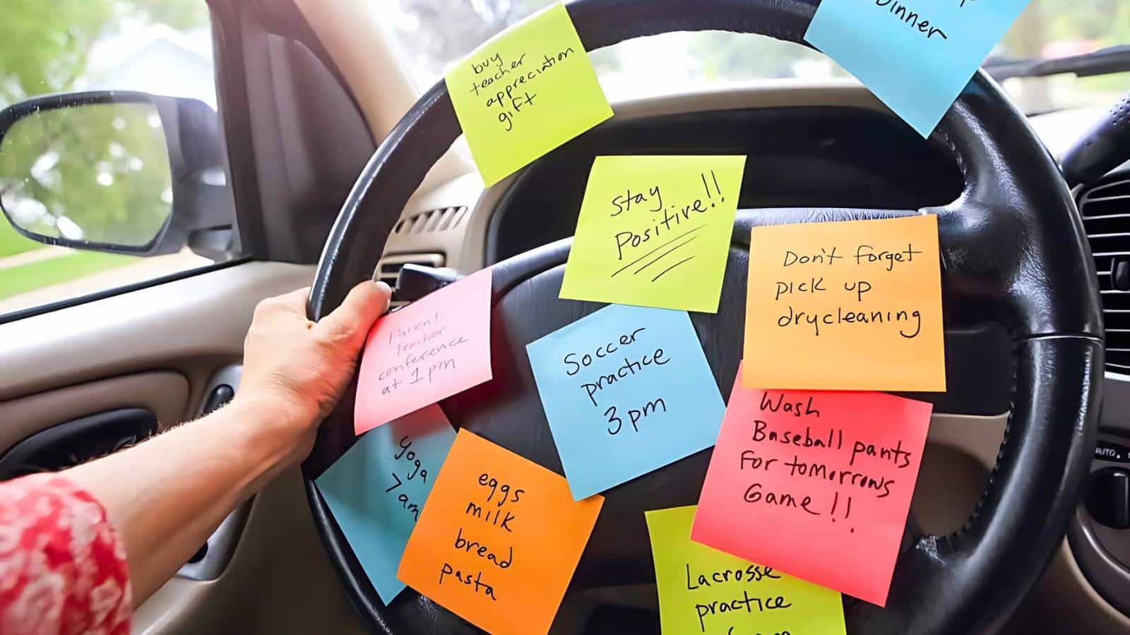 Steering wheel covered with colorful sticky notes, each listing various reminders such as grocery items, appointments, and motivational phrases. A hand is visible gripping the wheel.