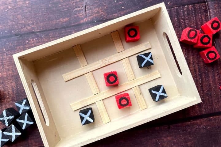 A wooden tic-tac-toe game board with red and black blocks marked with X and O. The game is in progress, with X blocking O in the central row. Additional blocks are scattered around the board.