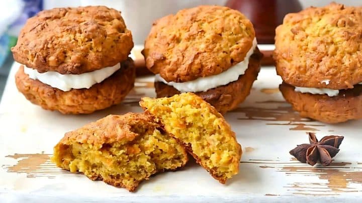 Four pumpkin cookies with cream filling are arranged on a wooden surface. One cookie is broken apart, revealing its texture. Anise stars are also visible.
