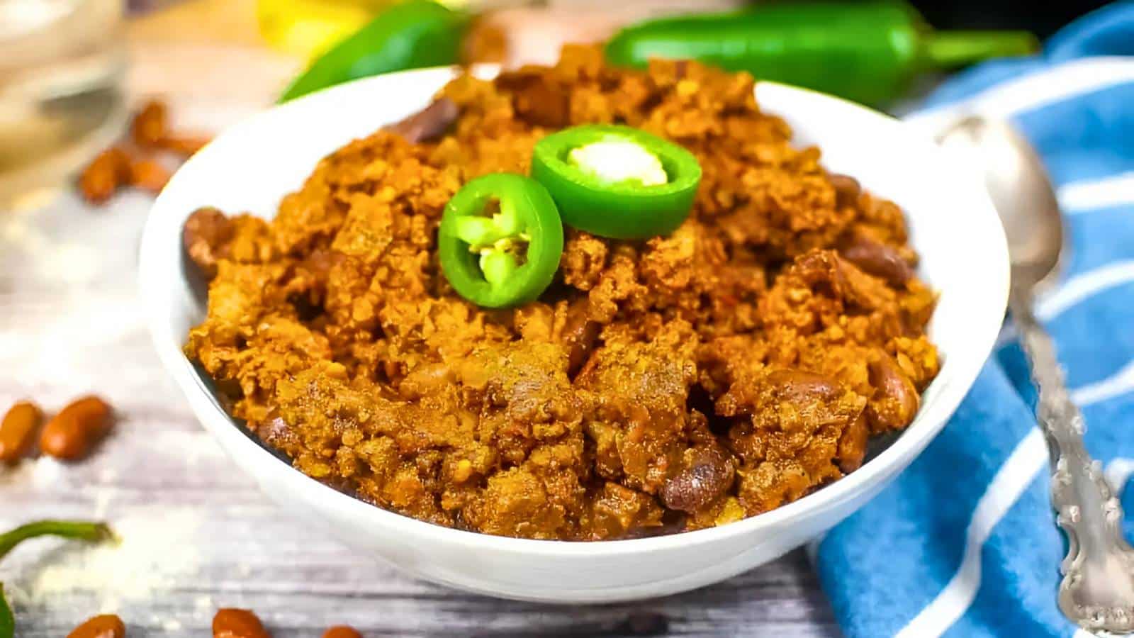 A white bowl filled with chili con carne, topped with sliced green chilies. A blue and white napkin and silver spoon are in the background.