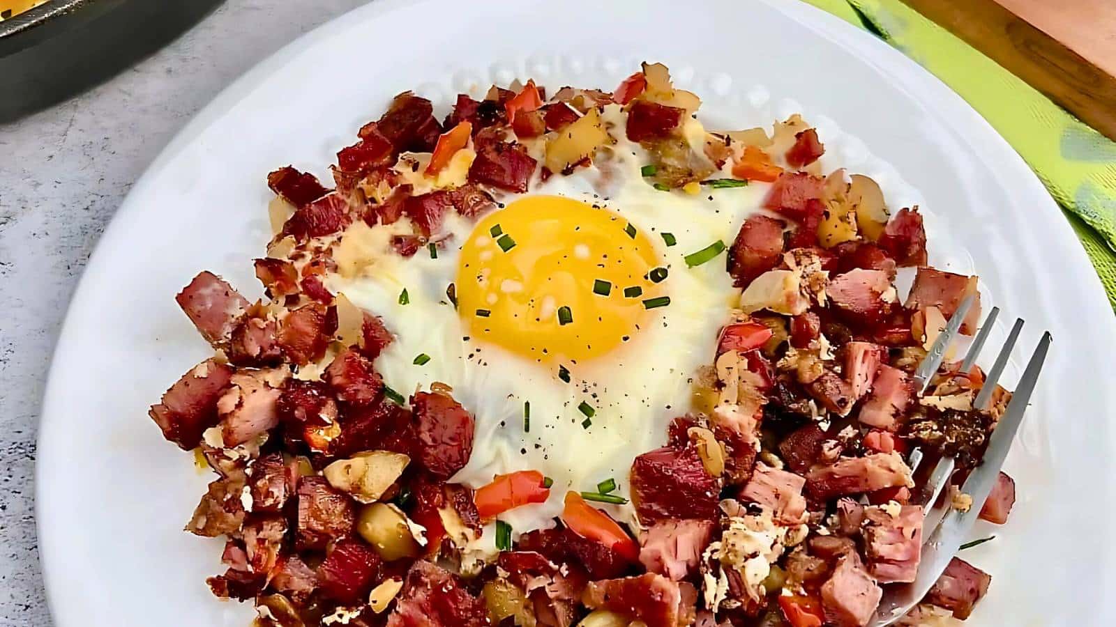 A plate of corned beef hash with diced vegetables and a sunny-side-up egg on top. A fork is placed on the side.