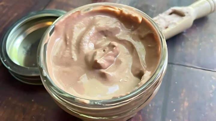 An open jar of thick, creamy chocolate spread sits on a wooden surface, with a spoon in the background.
