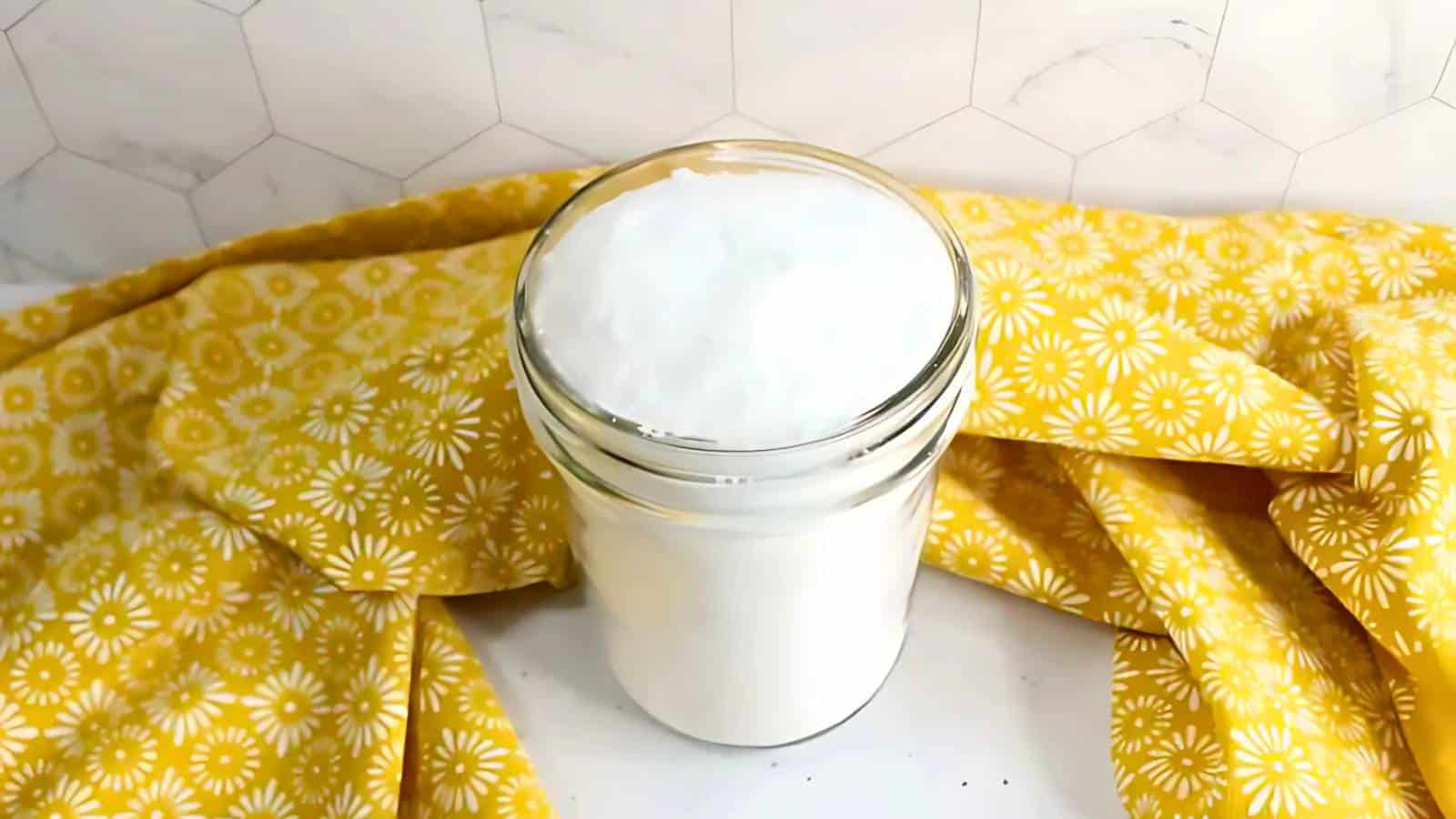 A glass jar filled with white powder is placed on a white surface. A yellow floral-patterned cloth surrounds the jar. A hexagonal-tiled wall is in the background.