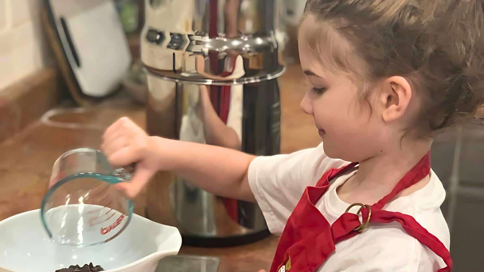 Child in a red apron pours ingredients from a measuring cup into a bowl on a kitchen counter.