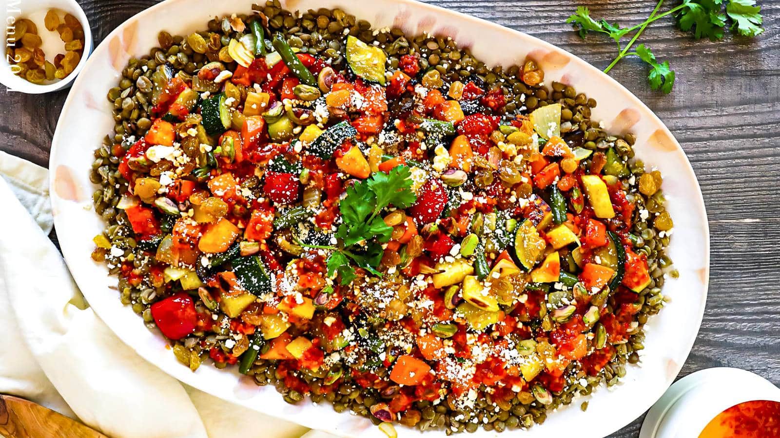 A large white dish contains a colorful lentil salad with chopped vegetables, herbs, and crumbled cheese, garnished with a sprig of fresh parsley.