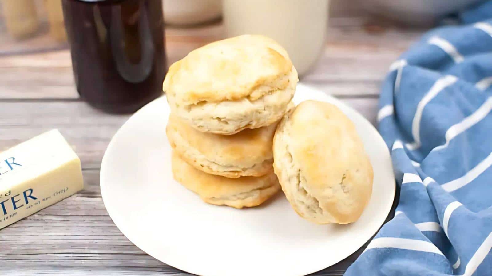 A plate with three golden-brown biscuits, straight from cozy sourdough recipes, sits on a wooden table next to a stick of butter and a blue-striped cloth.