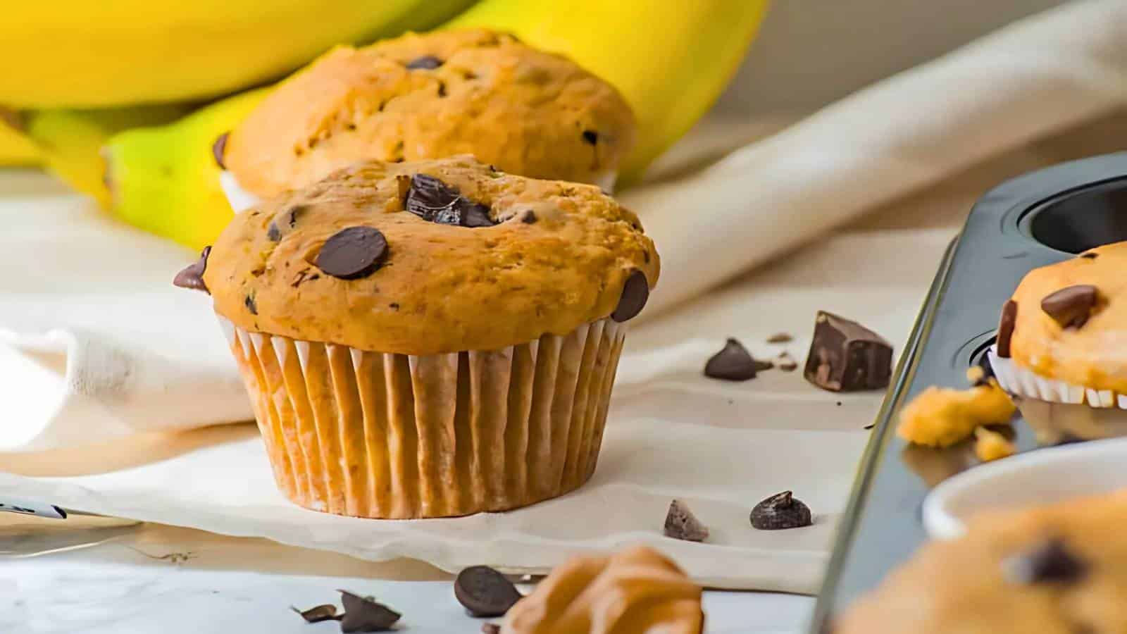 A chocolate chip muffin on a table with banana and chocolate pieces in the background, next to a muffin tin.