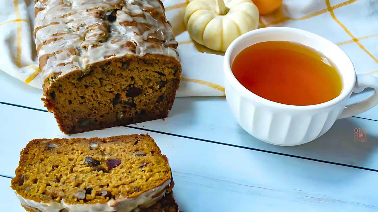A loaf of glazed pumpkin bread with two slices cut, next to a white cup filled with tea or coffee, with small pumpkins in the background.