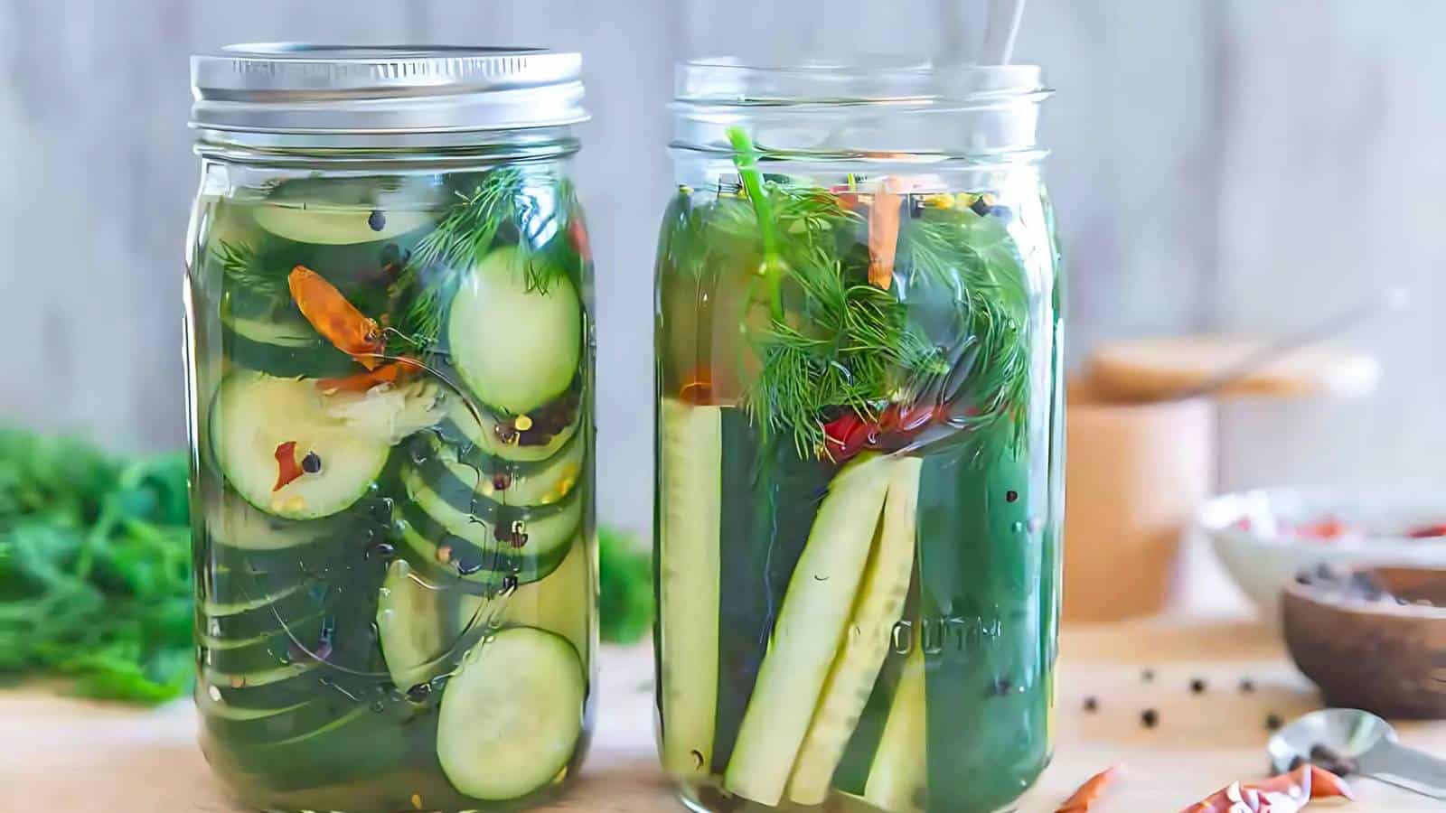Two jars of homemade pickles with sliced cucumbers, herbs, and spices are placed on a wooden surface with seasoning ingredients in the background.