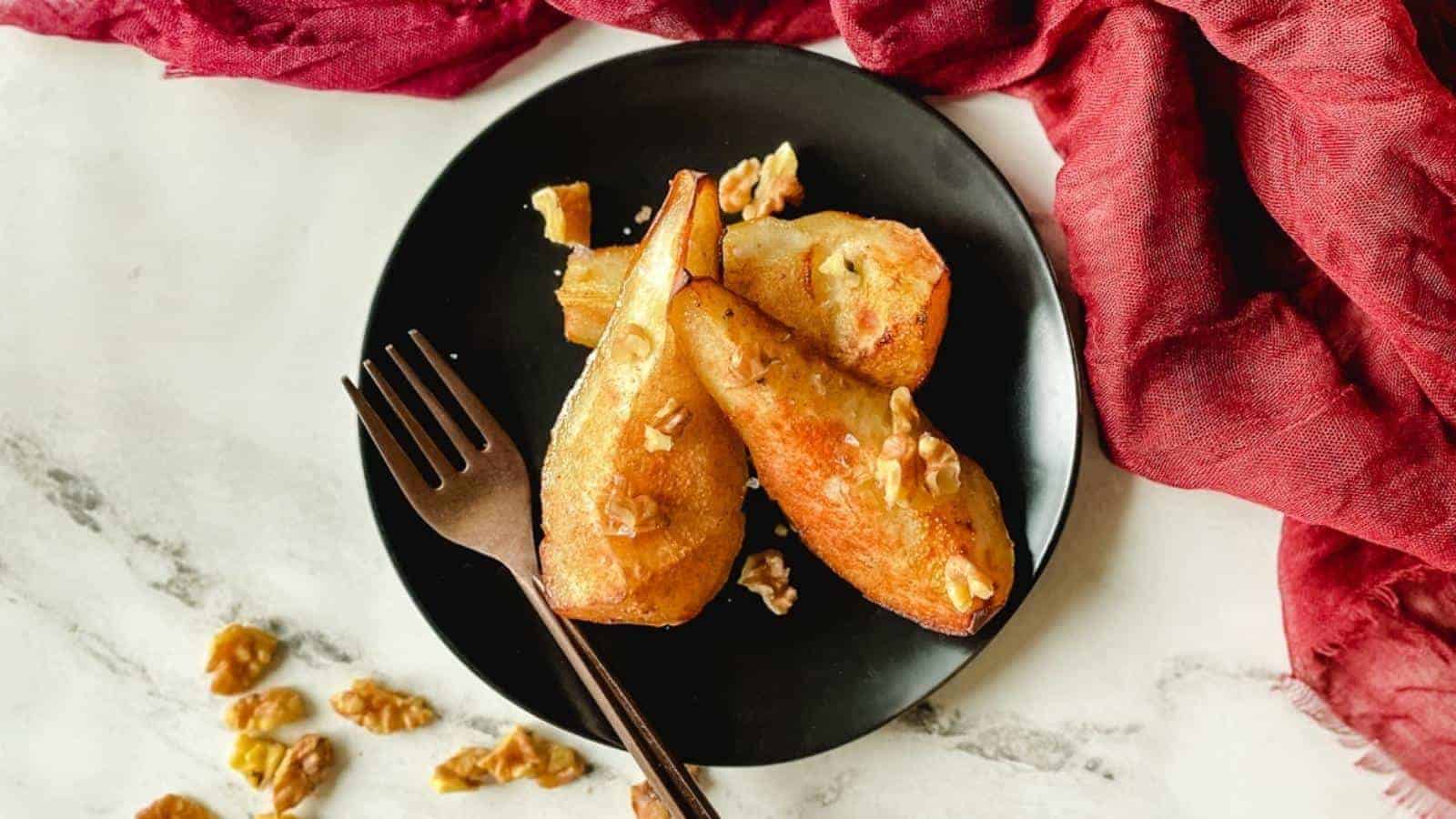 Grilled pear slices on a black plate, garnished with walnuts. A fork is placed beside them. A red cloth drapes nearby on a marble surface.