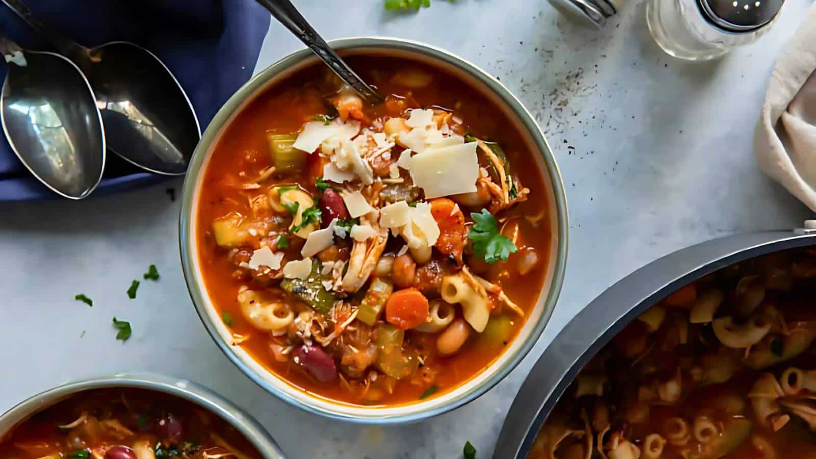 A bowl of vegetable soup with pasta, garnished with parsley and shaved cheese. Spoons and a napkin are visible nearby.