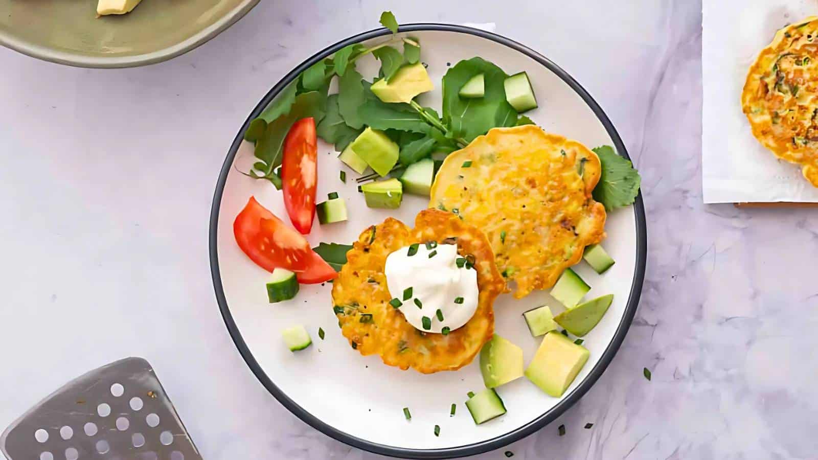 A plate with corn fritters topped with sour cream, garnished with chives. Diced avocado, sliced tomato, and greens are on the side.