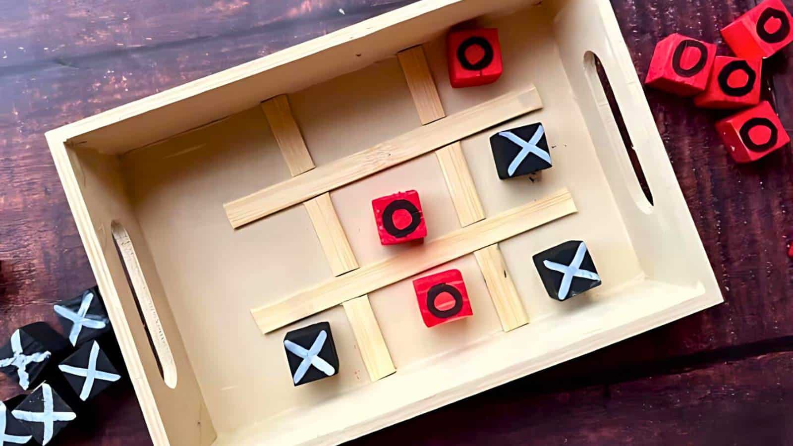 A wooden tic-tac-toe board with a grid made of wooden strips. Red and black blocks with Xs and Os are placed in the grid; red O wins diagonally. Extra blocks are on the side.