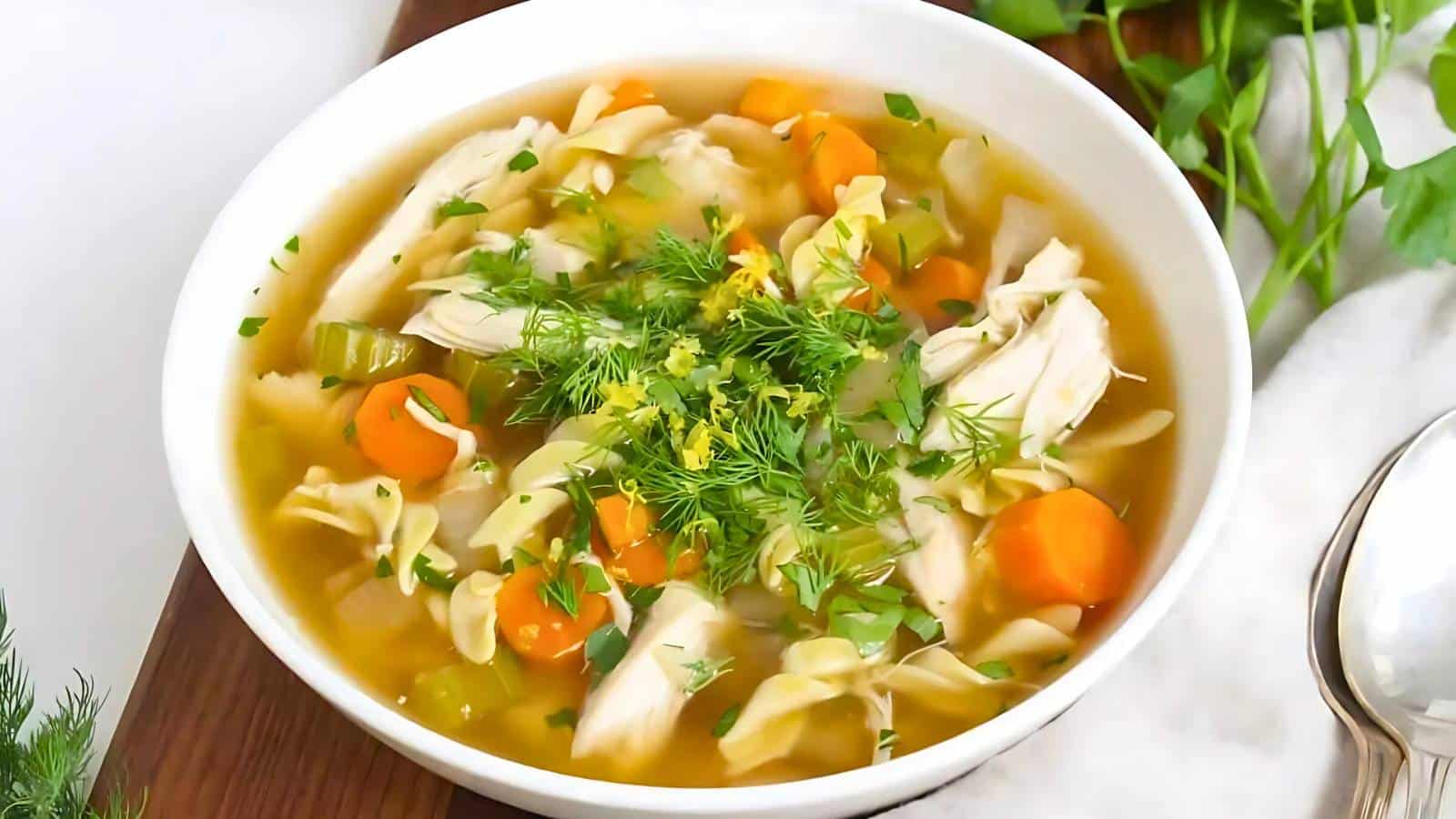 Two bowls of chicken noodle soup with vegetables and herbs, placed on a wooden board with parsley and two spoons nearby.