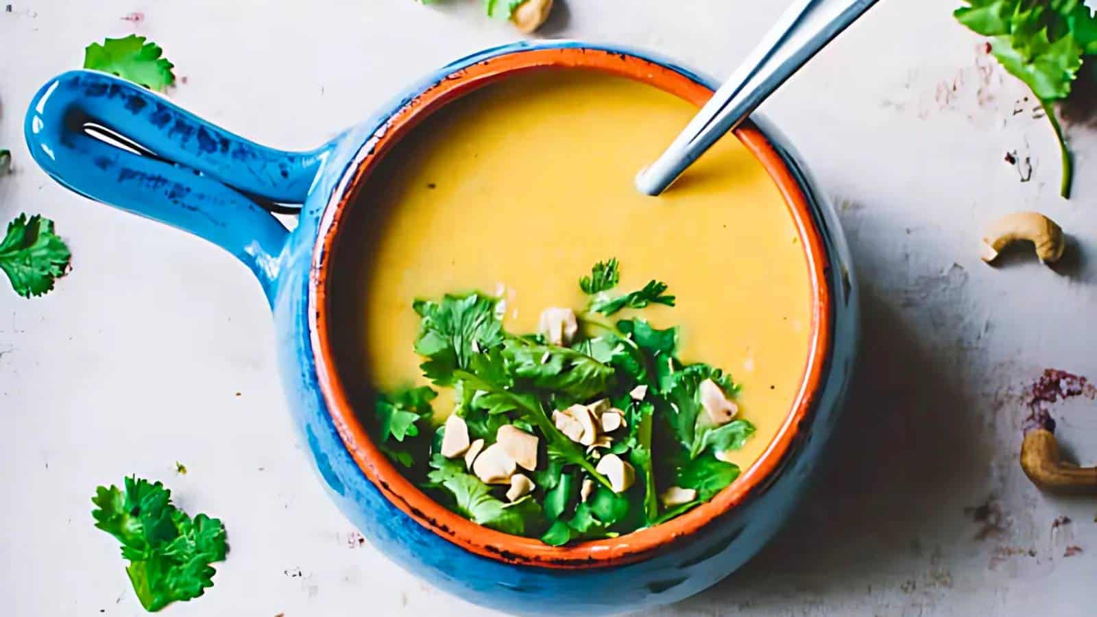 A blue bowl of creamy soup garnished with cilantro and cashews, with a spoon inside.