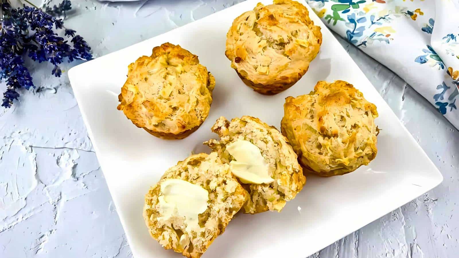 Four savory muffins are arranged on a white plate. One muffin is cut open, topped with melting butter. A floral cloth is in the background.