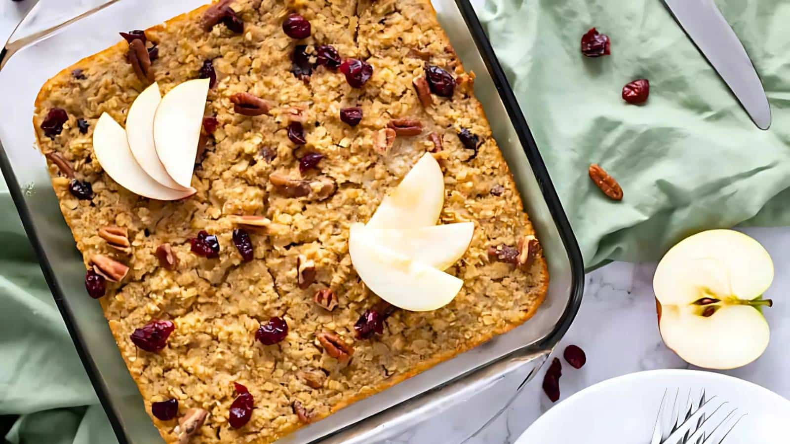 Baked oatmeal with cranberries and pecans in a glass dish, garnished with apple slices. Green cloth and apple halves nearby.