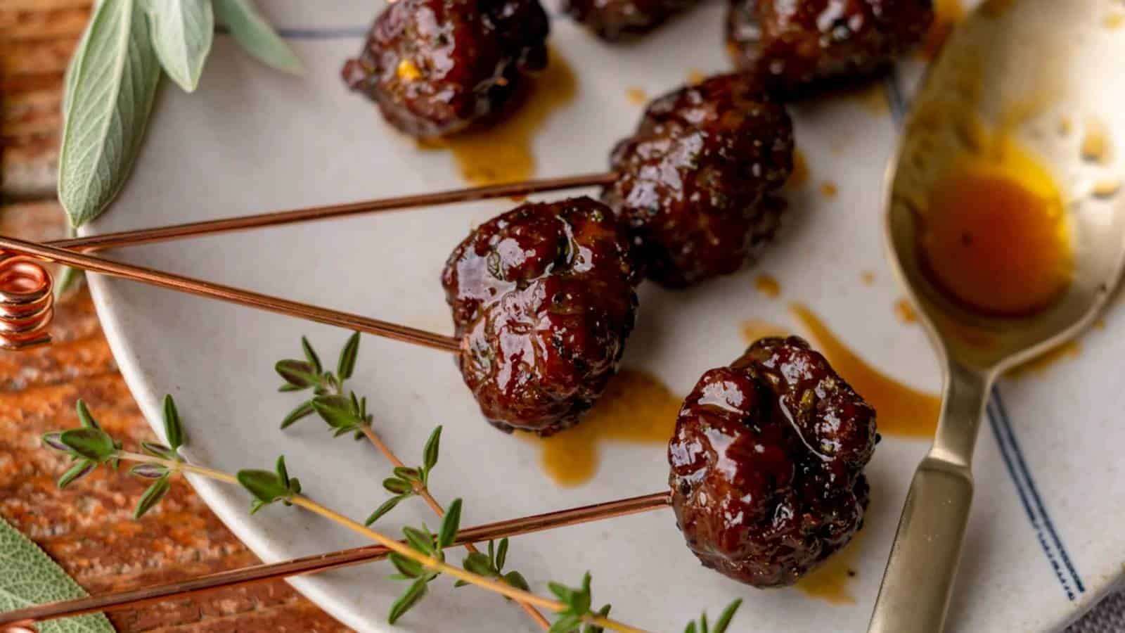 Skewered glazed meatballs on a plate with herbs and a spoon with sauce.