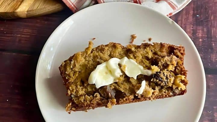 A slice of banana bread with a dollop of cream rests on a white plate, exuding cozy vibes perfect for hearty fall breakfasts. It's set on a wooden table, with a napkin and cutting board partially visible, inviting you to savor every moment.