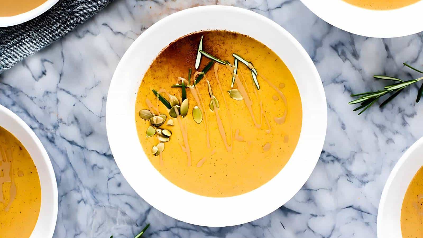 Bowls of creamy orange soup garnished with pumpkin seeds and rosemary on a marble surface.